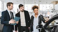 An interracial couple consults with a salesman at a car dealership, exploring vehicle options.